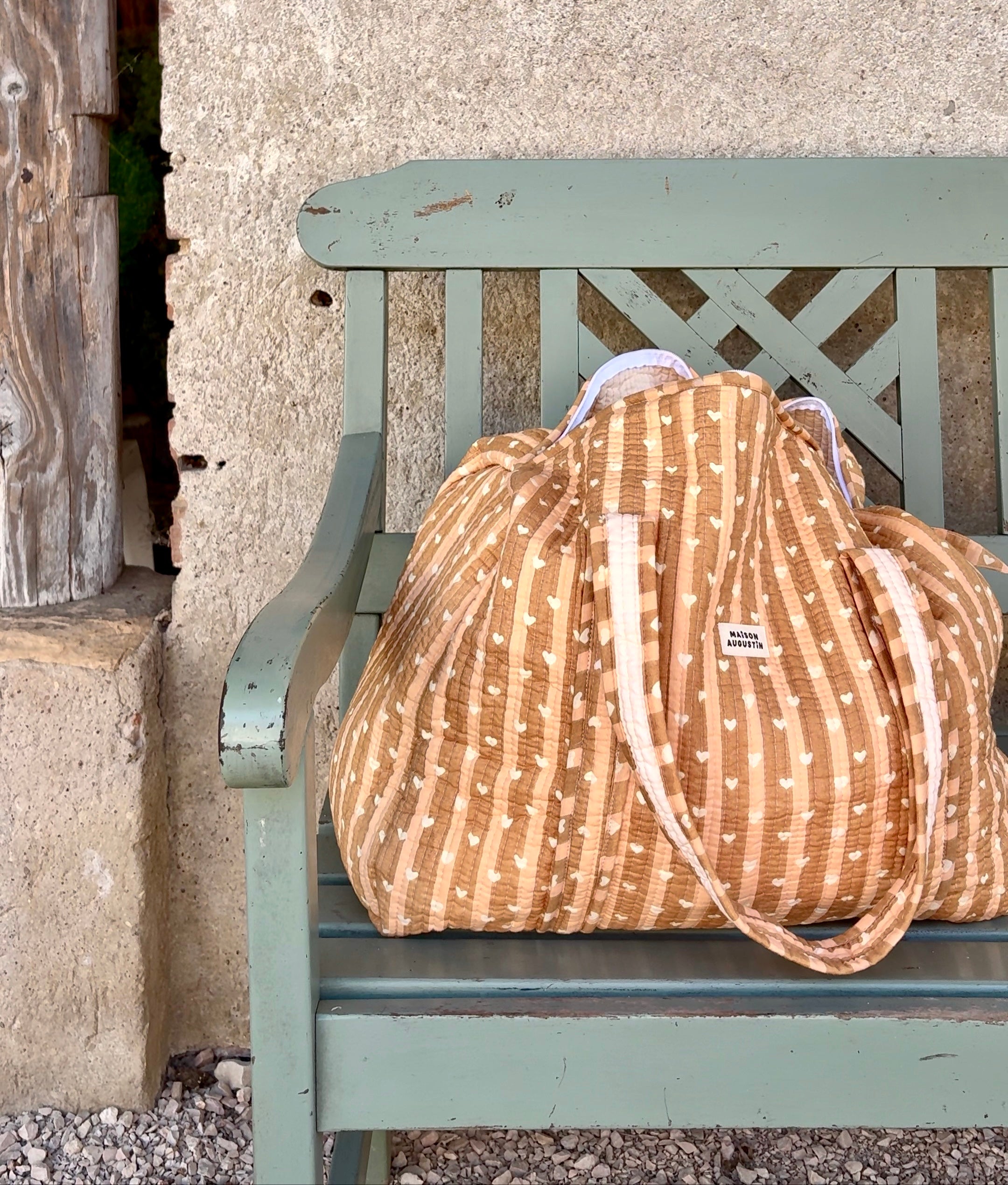 Sac week-end maternité cœur love beige  maison Augustin Posé sur un banc en bois vert d’eau 